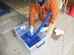 TGパウダー植毛塗装のモニター作業 接着剤を刷毛で満遍なく塗り広げます。かなり明るい環境で行わないと塗りムラの原因となります。接着剤が少ないと最後のエアブローで悲惨な状況を迎えてしまうため、しっかりと塗ります。その後、適当なボックスの中で粉を満遍なく振りかけていきます。理想を言えば対象物が地面に置けるくらいボックスに余裕があったほうが良いです。写真のように手に持って作業をすると、思わぬところに素材をぶつけてTGパウダーが寄ってしまうこともあります。