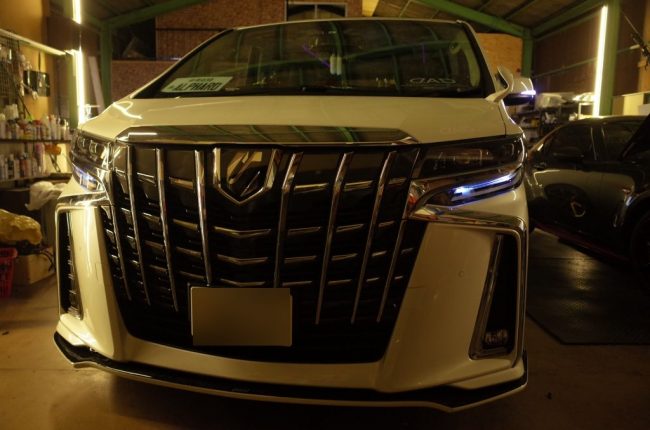 明るいガレージ内で、カスタム後のヘッドライト(デイライト点灯)が美しく輝くホワイトの30アルファード後期型のフロント全体像。
