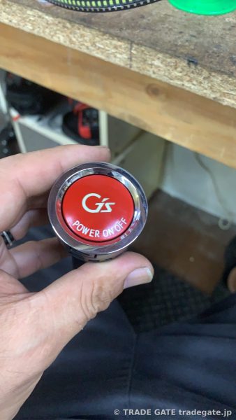作業員の手のひらに乗せられた、G'sロゴ入りの赤いプッシュスタートボタンのアップ。分解された部品であり、打ち替え作業前の状態を示す。